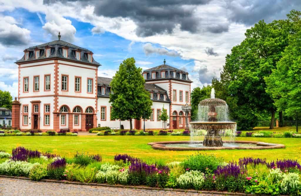 Elegantes Gebäude mit Springbrunnen in Hanau, von üppigem Grün und farbenfrohen Blumen umgeben, ideal für Immobilienmakler Hanau, die luxuriöse Immobilien präsentieren.