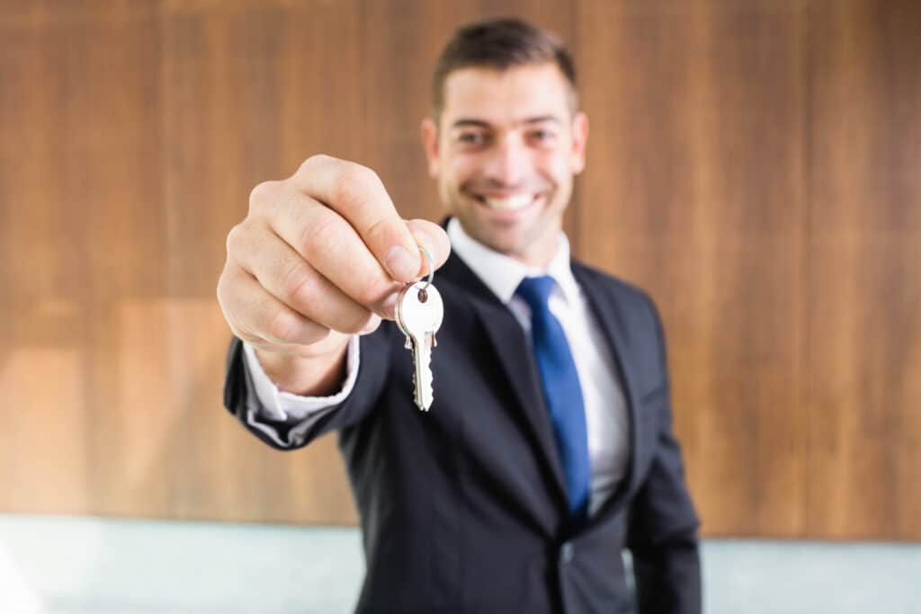 Strahlender Immobilienmakler aus Hofheim reicht Schlüssel, im Hintergrund unscharfes Bürointerieur.