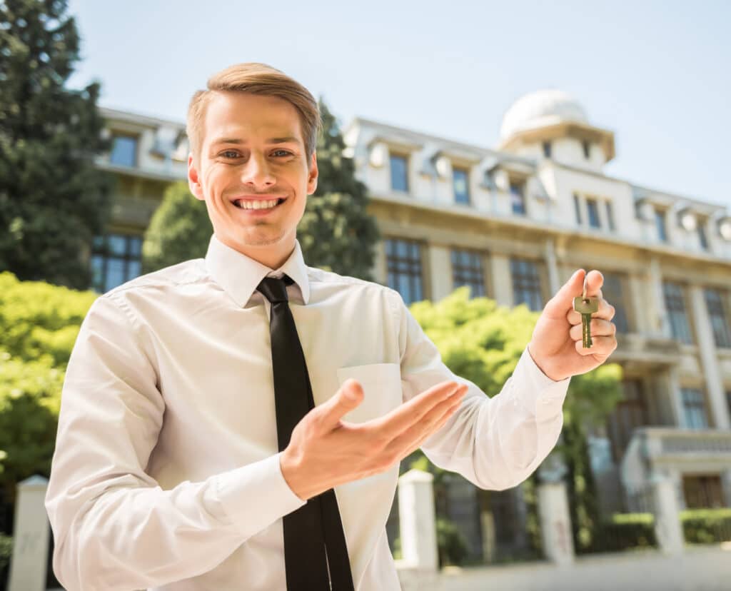 Lächelnder Immobilienmakler in weißem Hemd und Krawatte präsentiert Schlüssel vor einem Gebäude, repräsentiert Qualität und Service in Weinheim.