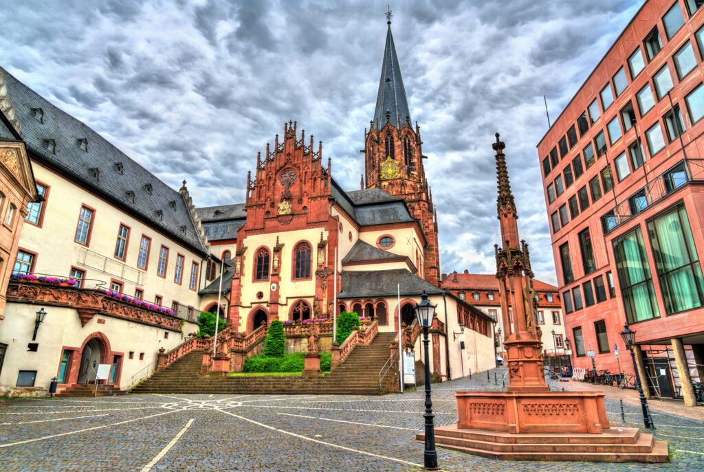 Die St. Peter und Alexander Kirche und der Abteibrunnen in Aschaffenburg, umgeben von historischen Gebäuden, die von unseren Immobilienmaklern in Aschaffenburg geschätzt werden.